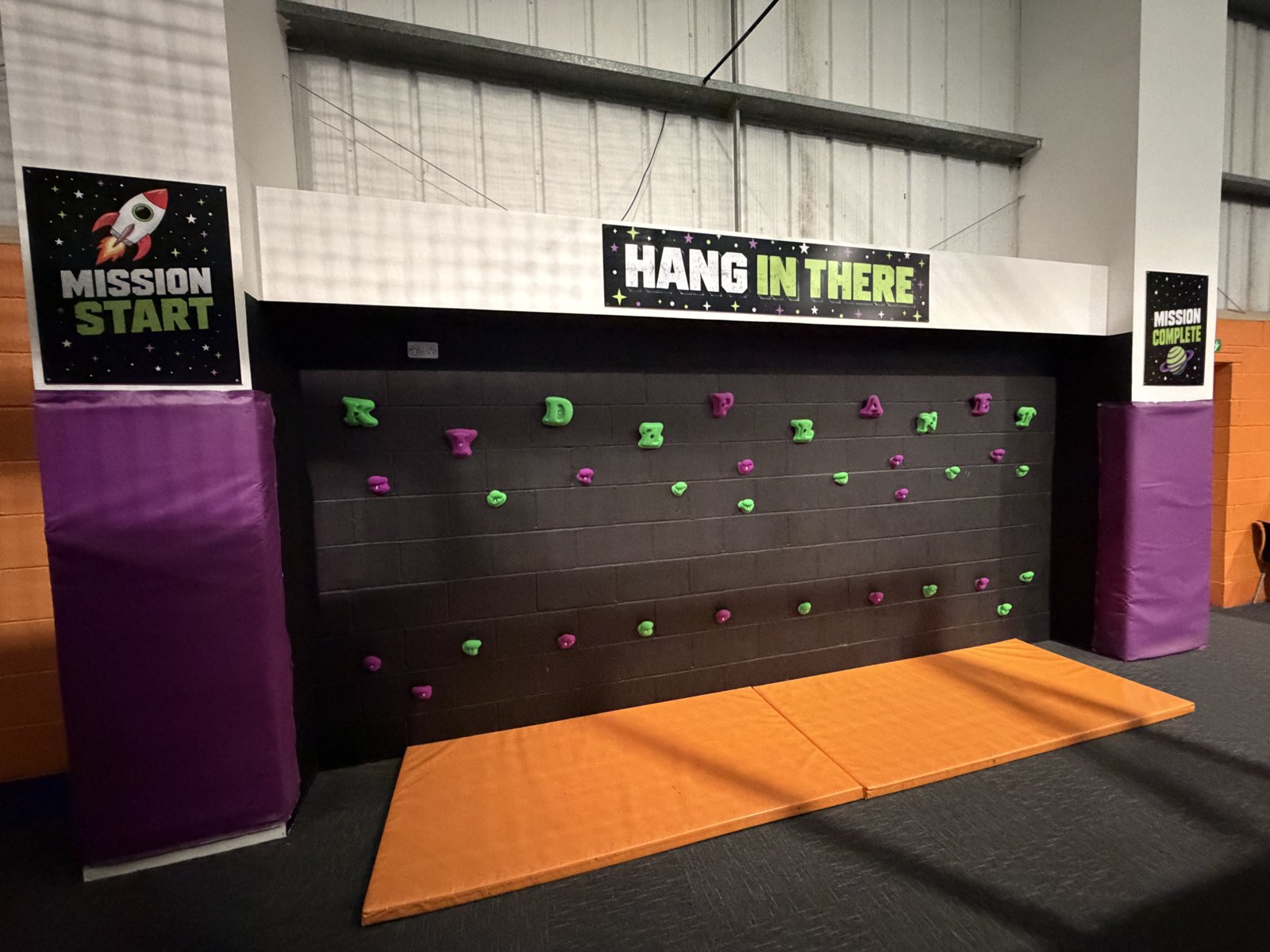 The Hang In There climbing wall with green and purple holds spelling Kidz Planet, Mission Start and Mission Complete signs and orange crash mats